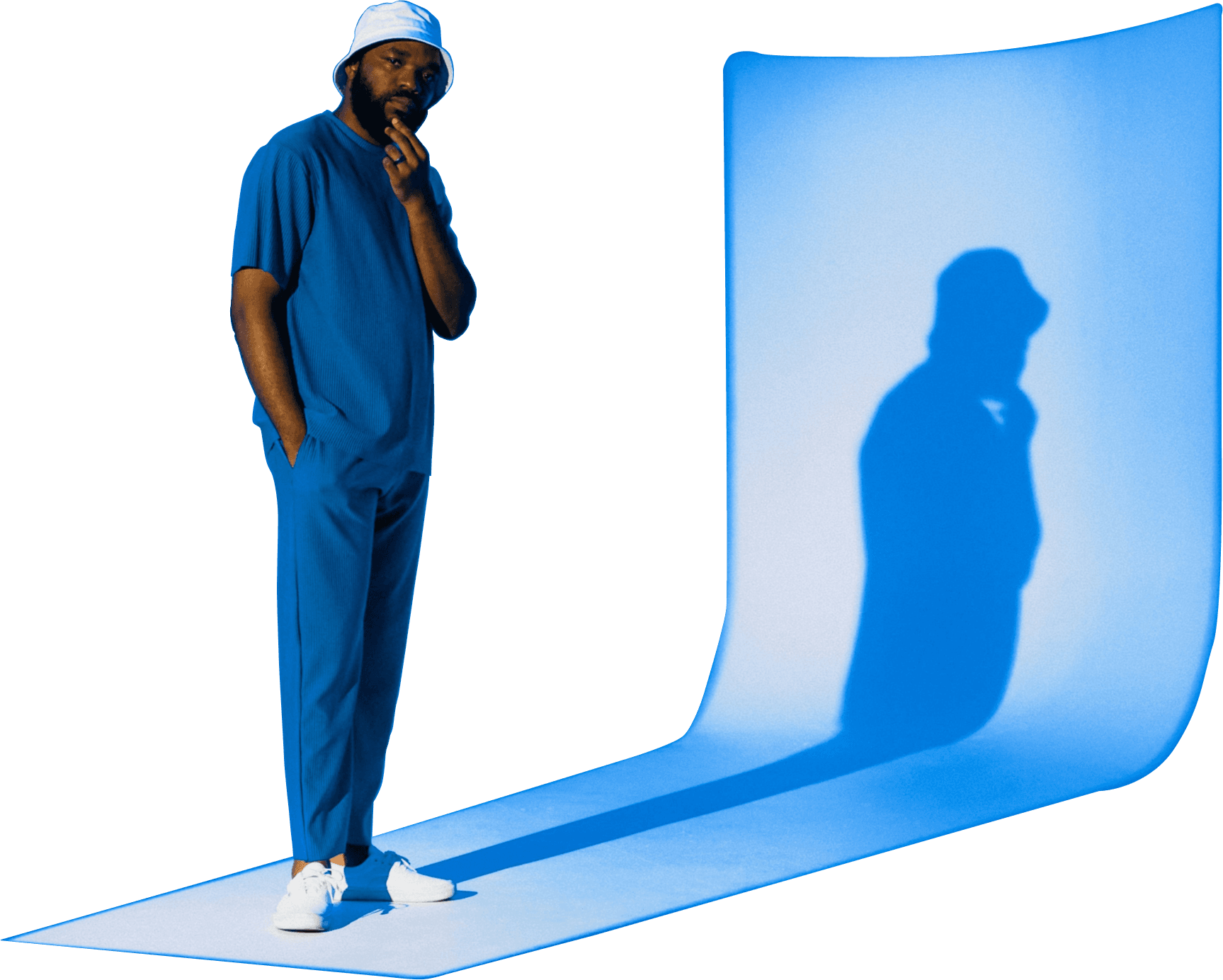 A man in blue clothing and a white bucket hat stands thoughtfully in a minimalist blue studio, casting a long shadow onto a curved backdrop lit by a spotlight